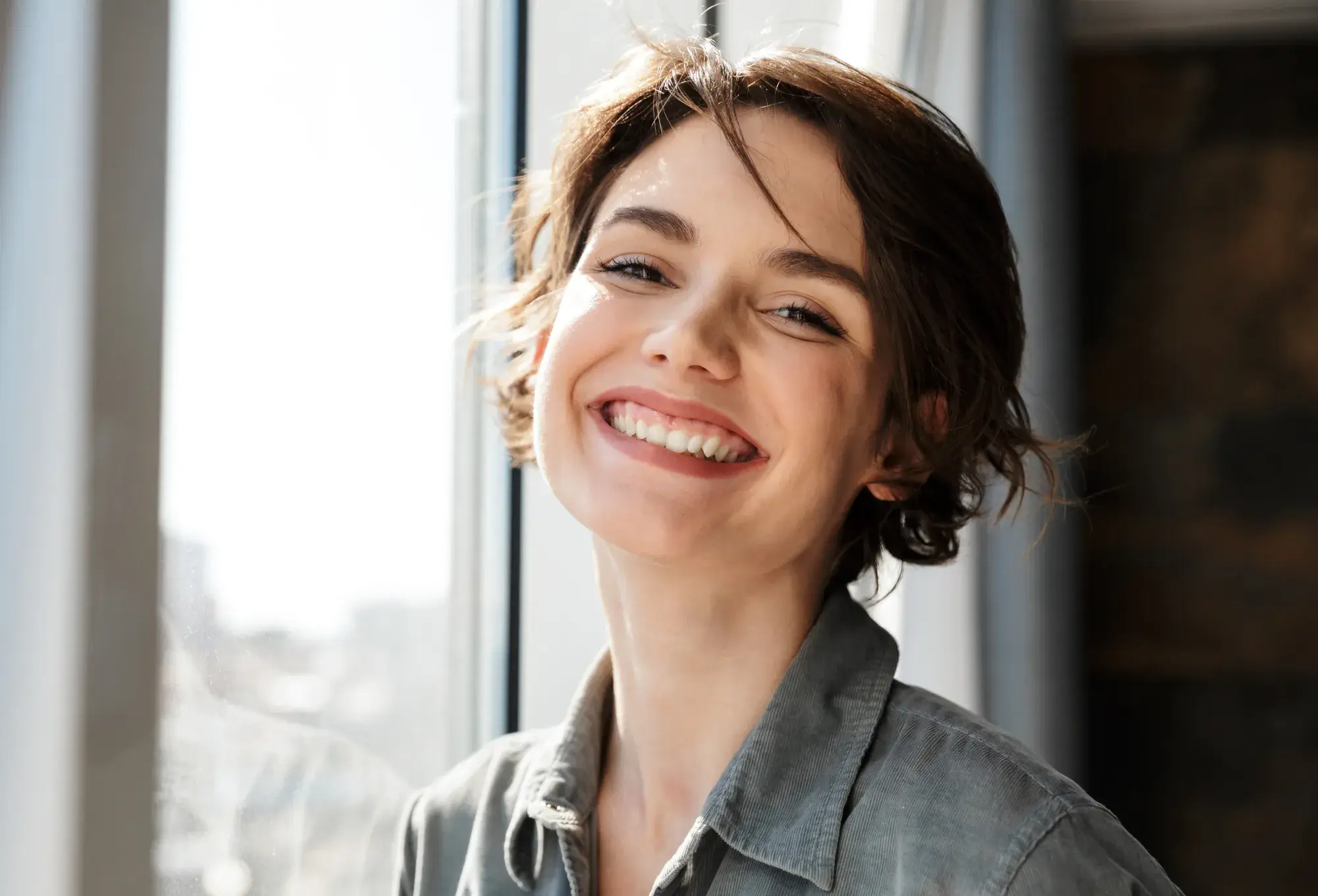 A photo of a smiling person with short hair, wearing a gray shirt, standing near a window with light coming in.