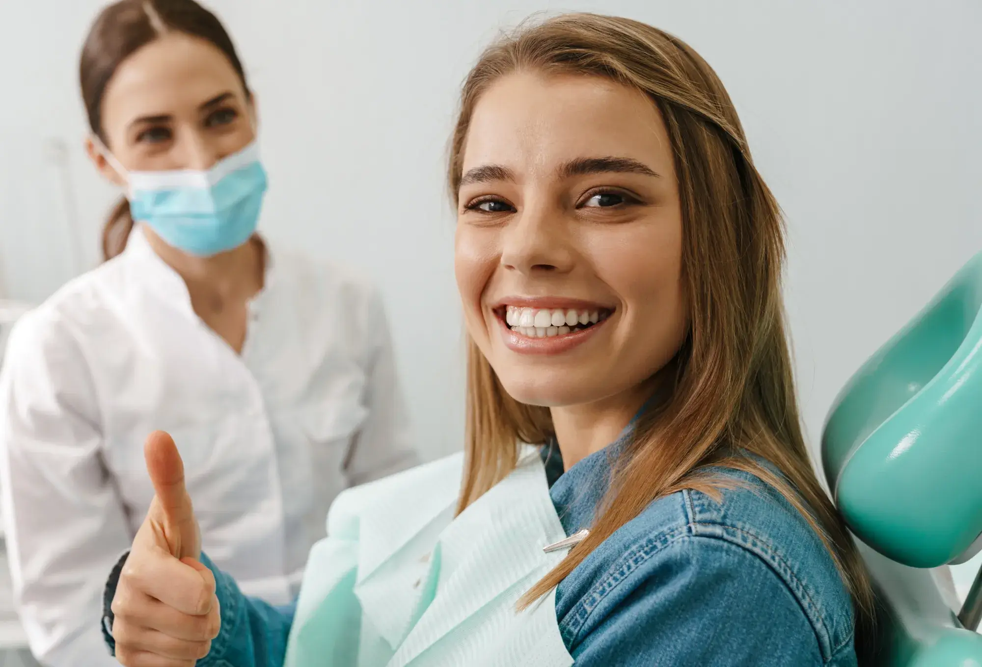 Photo of a woman in a dental chair smiling and giving a thumbs up, with a dentist in a mask beside her.