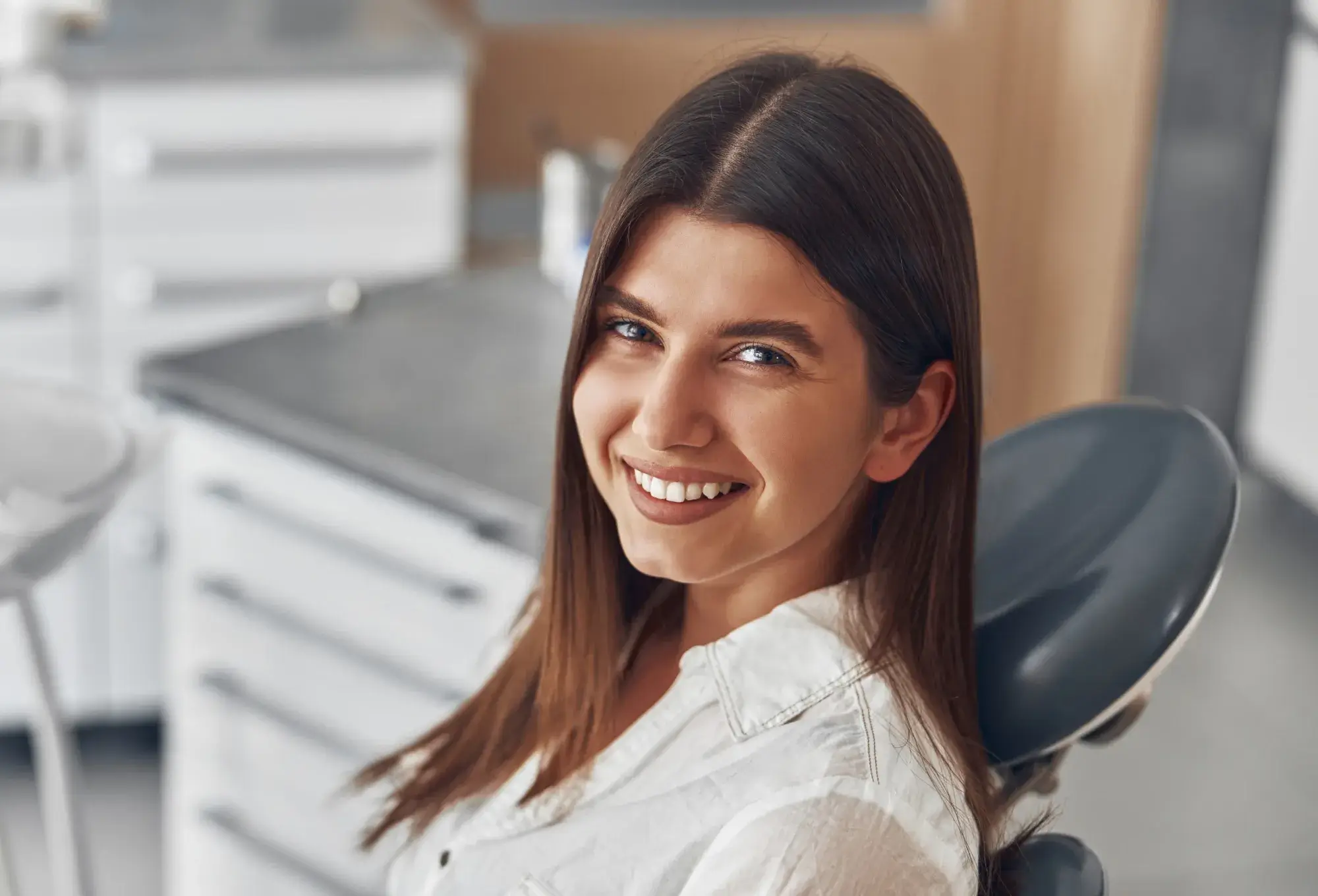 A photo of a smiling woman with brown hair sitting in a dental chair, wearing a white shirt.