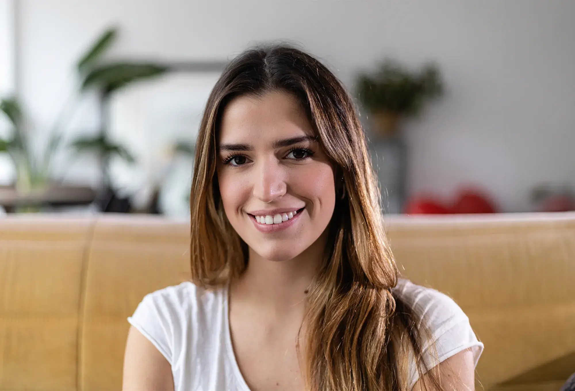A woman with long brown hair smiling indoors, sitting on a beige couch, plants in the background.