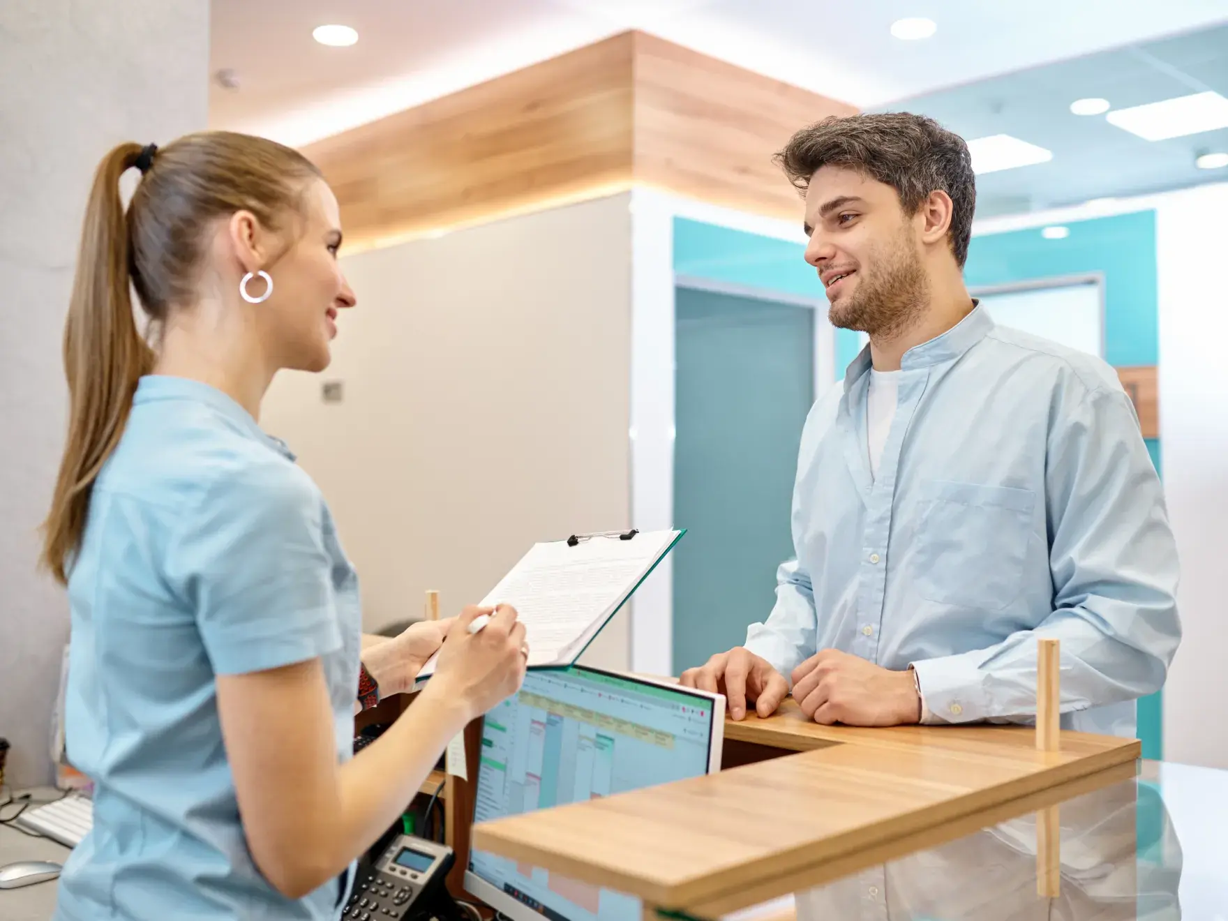Photo of a woman and a man at a reception desk talking, both wearing light blue uniforms.