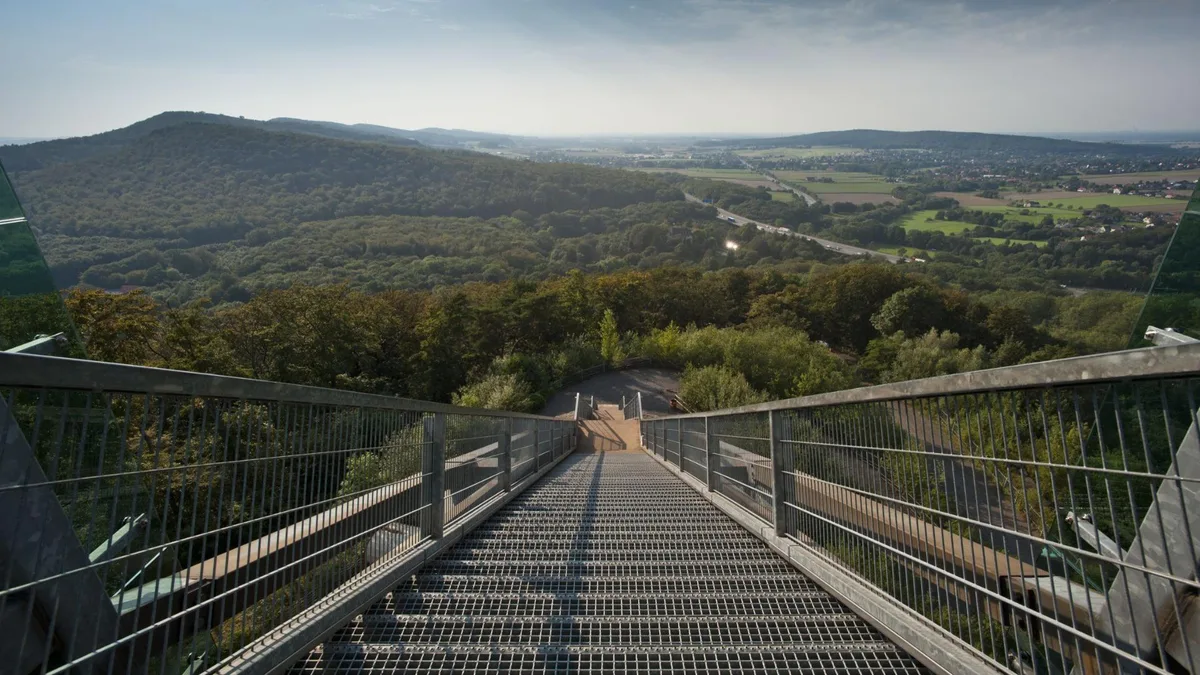 Gitterrost-Steg mit Geländer führt vom Aussichtsbauwerk abwärts, dahinter bewaldete Höhenzüge über dem Wesertal
