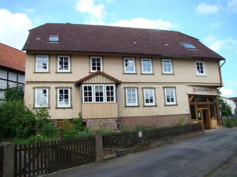 Zweigeschossiges Fachwerkhaus mit hellem Bretterverschlag und Sprossenfenstern an einer Dorfstraße, rechts der Eingang des Keramikmuseums unter einem Vordach, davor ein niedriger Lattenzaun