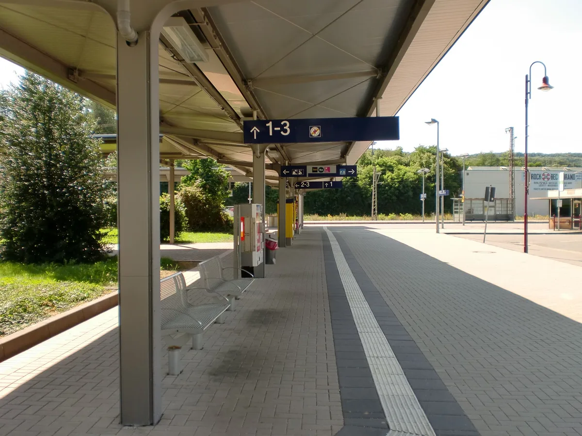 Überdachter Bahnsteig eines Regionalbahnhofs mit Anzeigetafel zu den Gleisen 1 bis 3, davor ein leeres Gleis