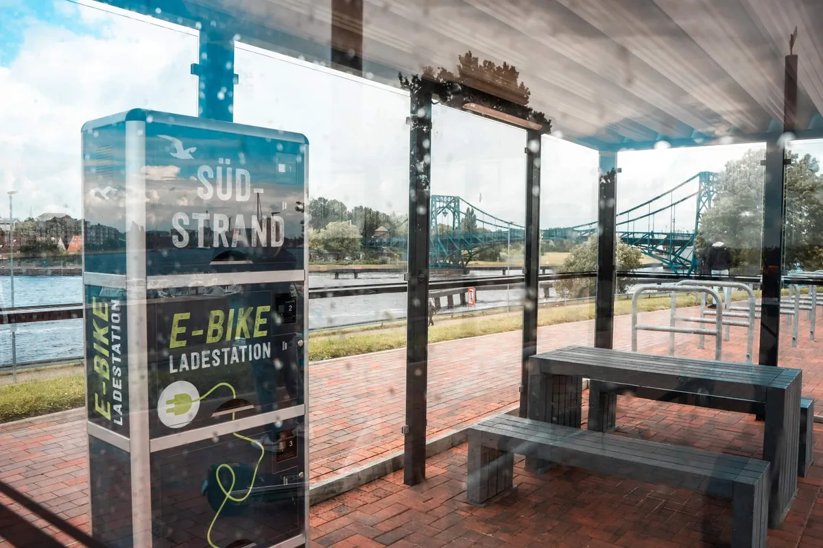 Überdachte Fahrradstation mit Sitzbänken und E-Bike-Ladestation am Südstrand, dahinter das Wasser der Jade und die Kaiser-Wilhelm-Brücke