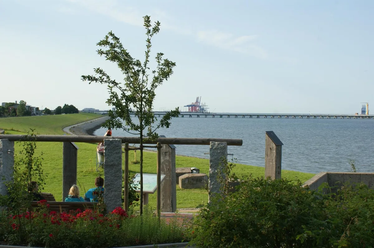 Rastplatz mit Bänken und Infotafel auf einer Deichwiese, dahinter der Jadebusen und eine Brücke am Horizont