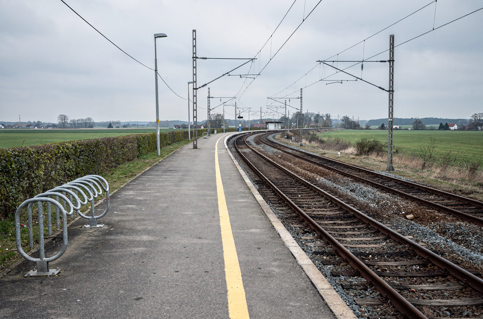KI-generiertes Bild: Leerer Bahnsteig auf dem Land mit gelbem Sicherheitsstreifen, links ein leerer Fahrradständer aus Stahlrohr und eine Hecke, rechts zwei Gleise, die in einer Kurve verschwinden, darüber Oberleitungsmasten