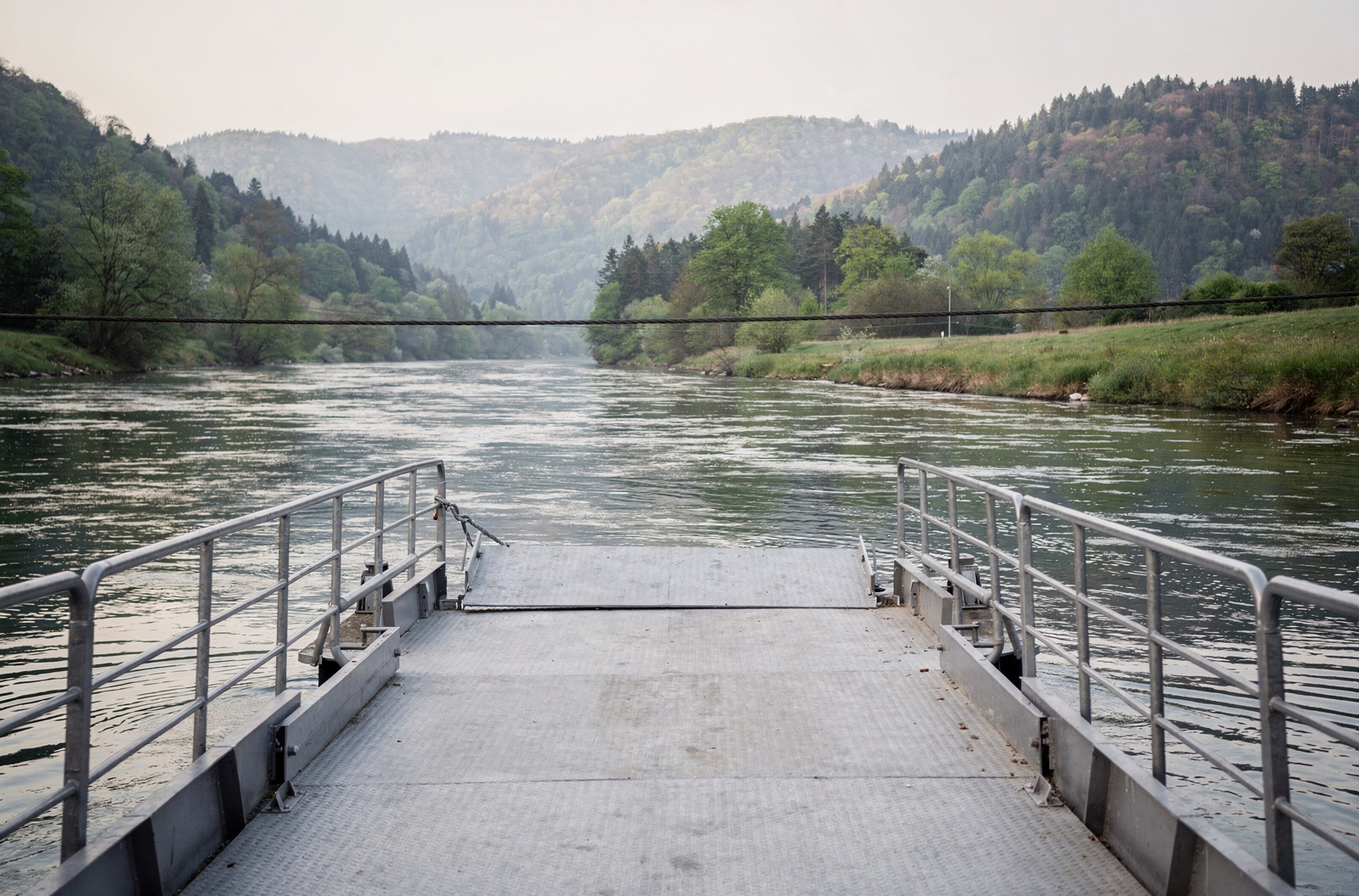 KI-generiertes Bild: Leeres Stahldeck einer Flussfähre mit heruntergelassener Rampe, davor ein Tragseil quer über den Fluss und bewaldete Hänge am Gegenufer