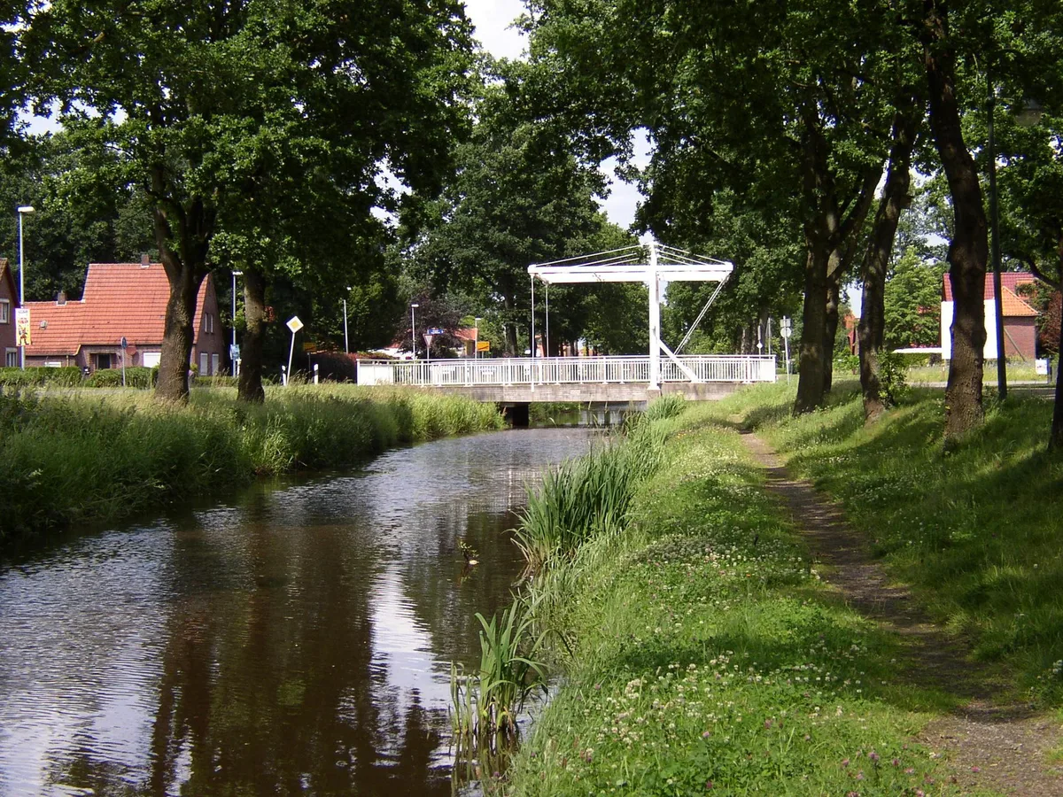 Ein Fehnkanal mit weißer Klappbrücke, daneben ein schmaler Treidelpfad unter Bäumen