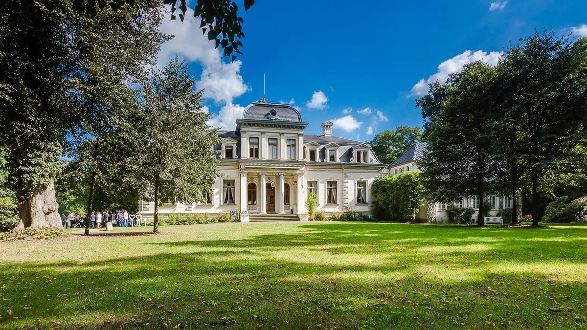 Das weiße Palais Rastede mit Säulenportikus und Mansarddach hinter einer weiten Rasenfläche, gerahmt von alten Laubbäumen, links eine Gruppe von Besuchern am Wegrand
