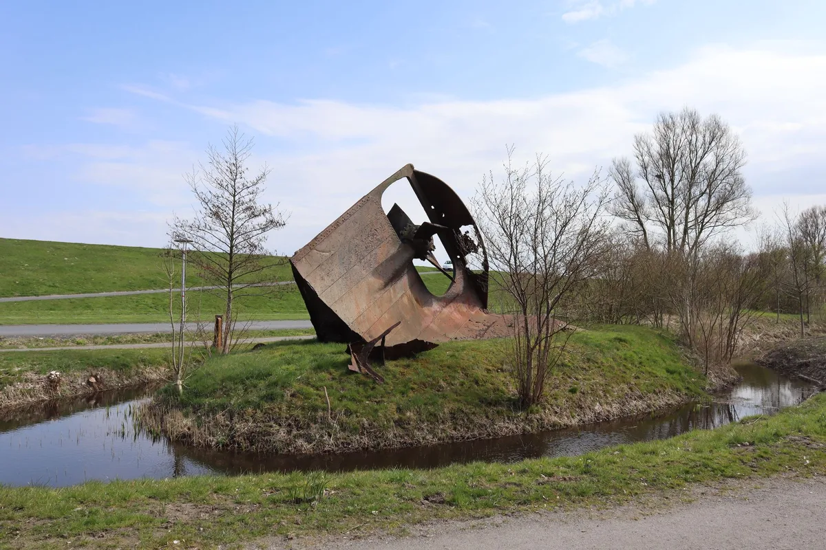 Rostrotes Wrackteil eines Schiffshecks auf einer Grasinsel am Otterndorfer Deich, ringsum ein Wassergraben, dahinter ein Weg und kahle Bäume.