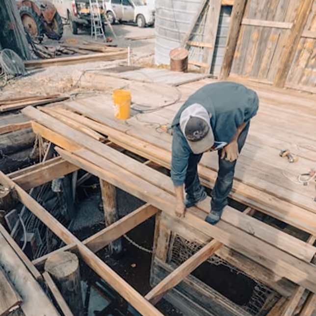 Construction worker wearing a cap and grey shirt bending over while working on a wooden deck's framework at a construction site with tools and vehicles in the background.