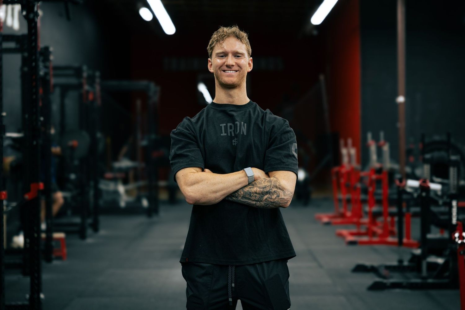 Ben Leonard smiling with arms crossed on the gym floor, red and black racks behind him