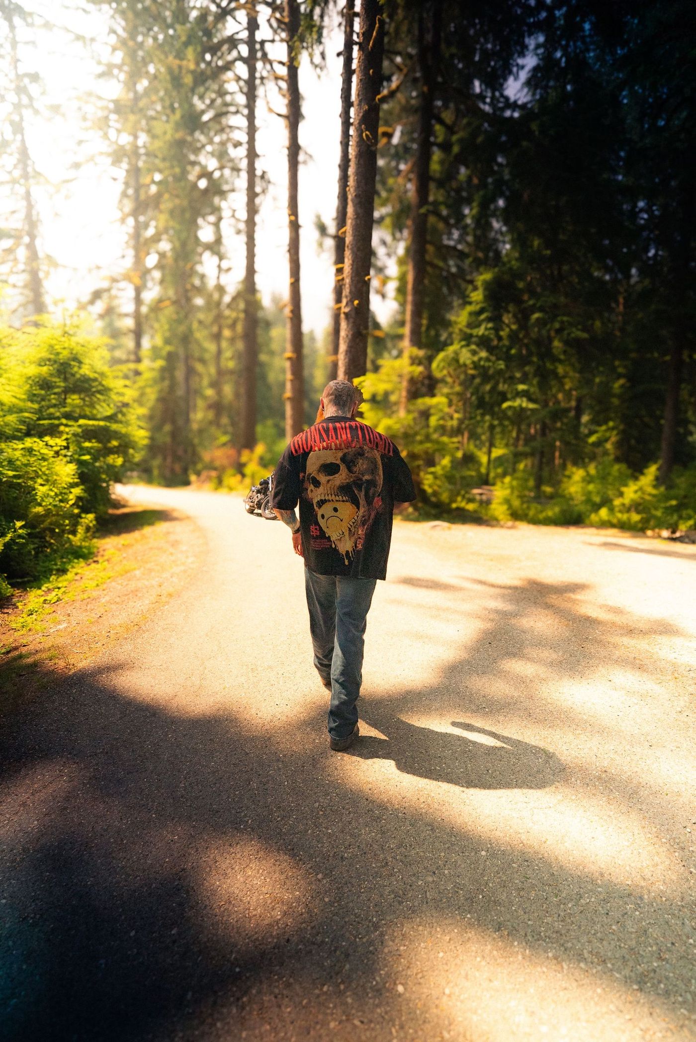 Walking away down a sunlit forest road in an Iron Sanctuary skull-back tee