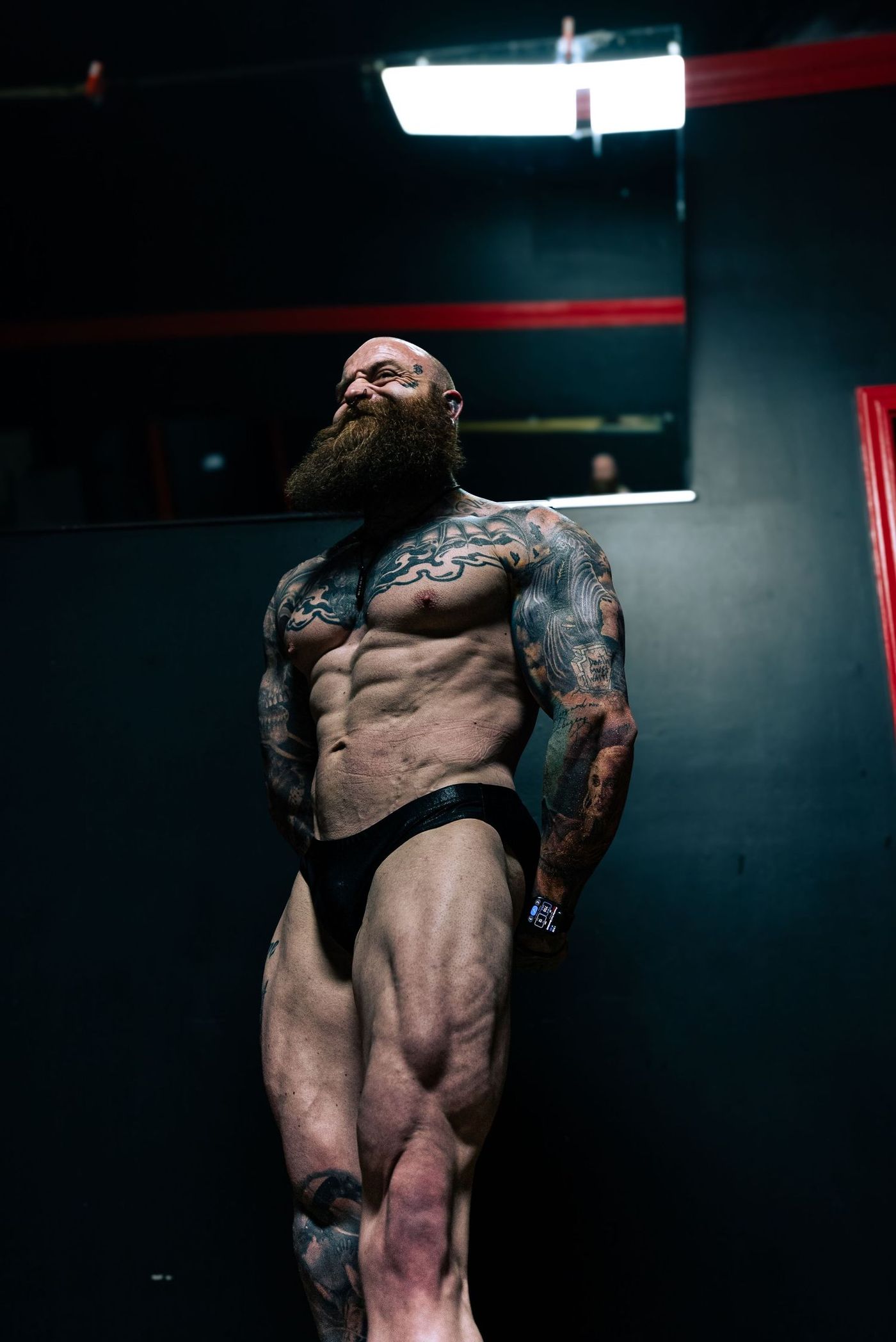 Bodybuilder holding a side pose under a single overhead light in a dark red and black room