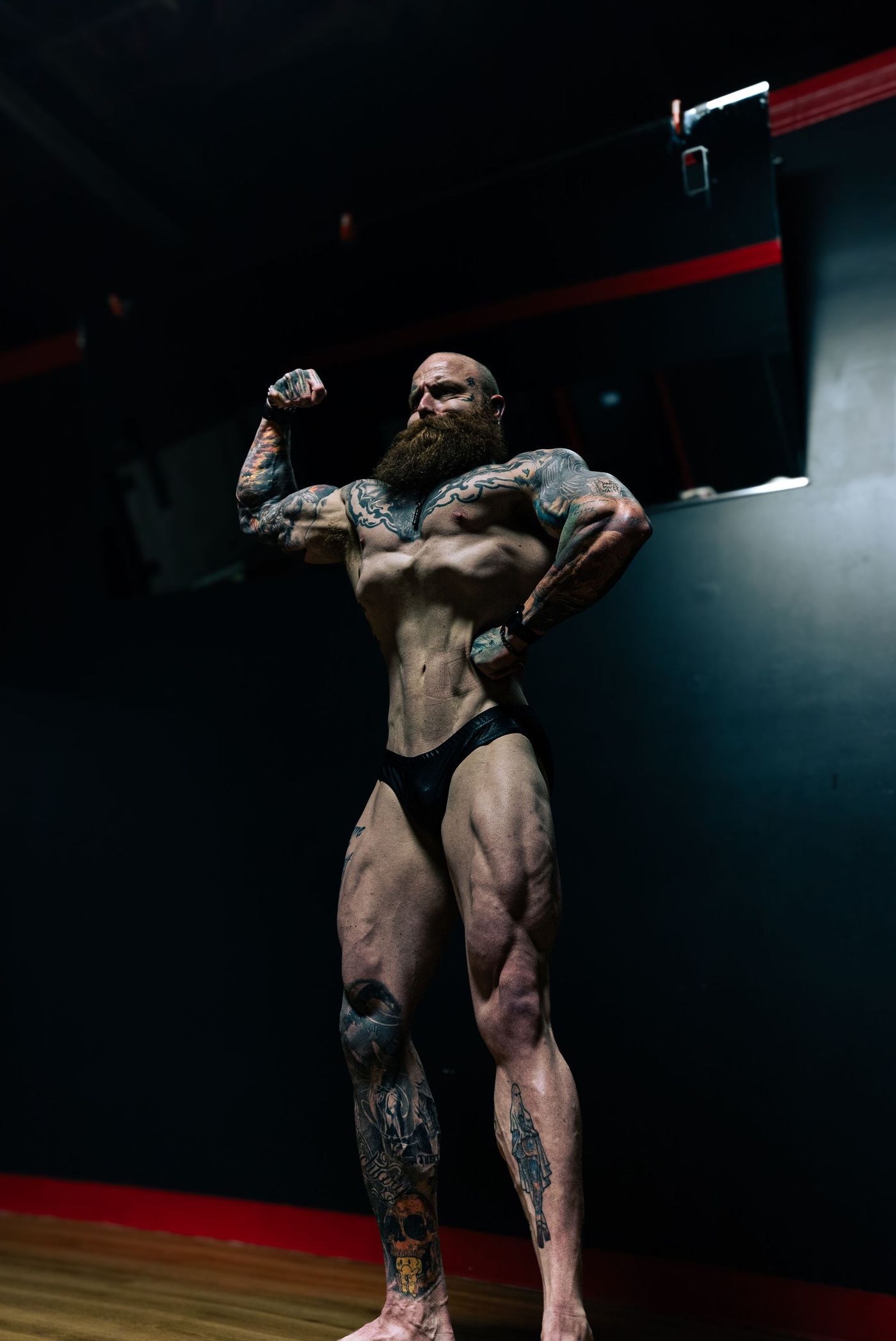 Bearded, tattooed bodybuilder hitting a front double bicep in posing trunks beside a red door frame