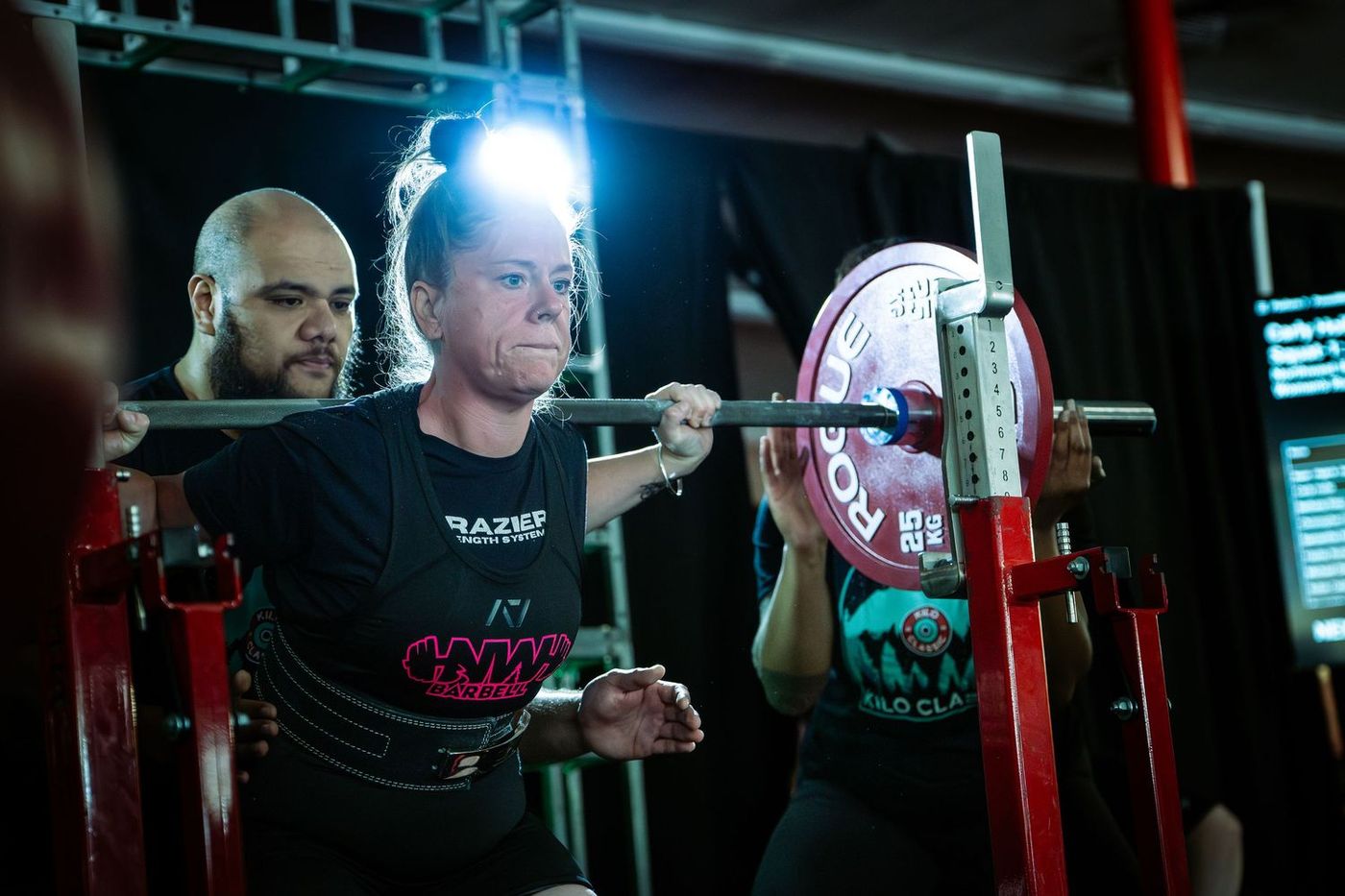 Lifter under the bar with a spotter behind her, red plates loaded, at a Powerlifting United meet
