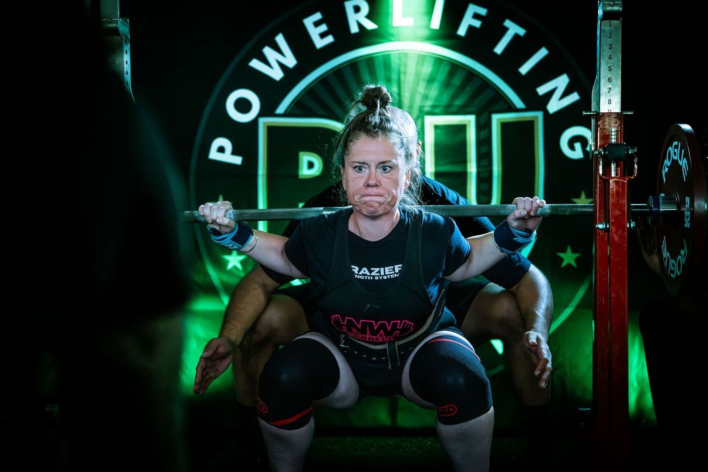 Lifter at the bottom of a squat in front of the green Powerlifting United logo, spotter behind her