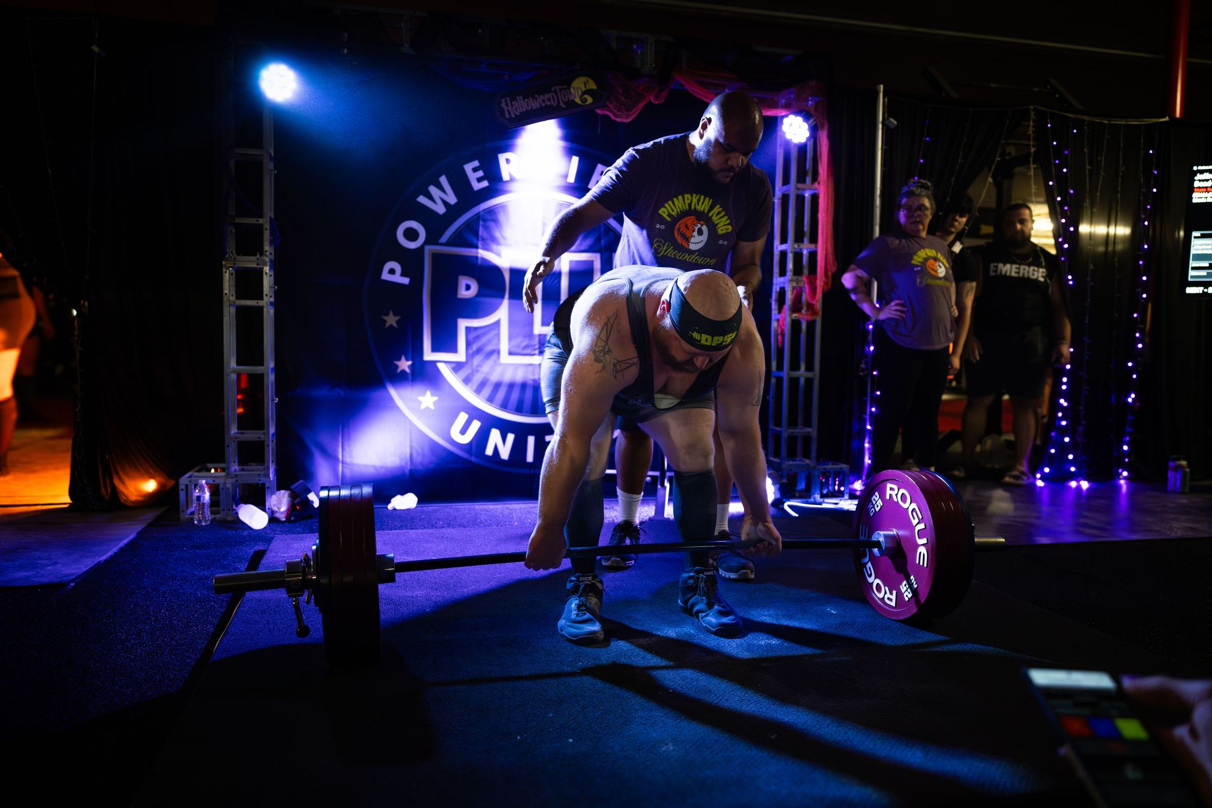 Lifter setting up a deadlift on a stage lit blue, Powerlifting United banner behind him