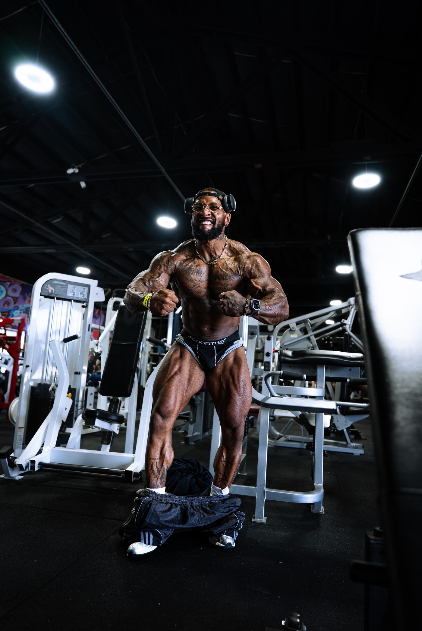 RIKFLEX holding a front pose in posing trunks on the gym floor, overhead lights behind him