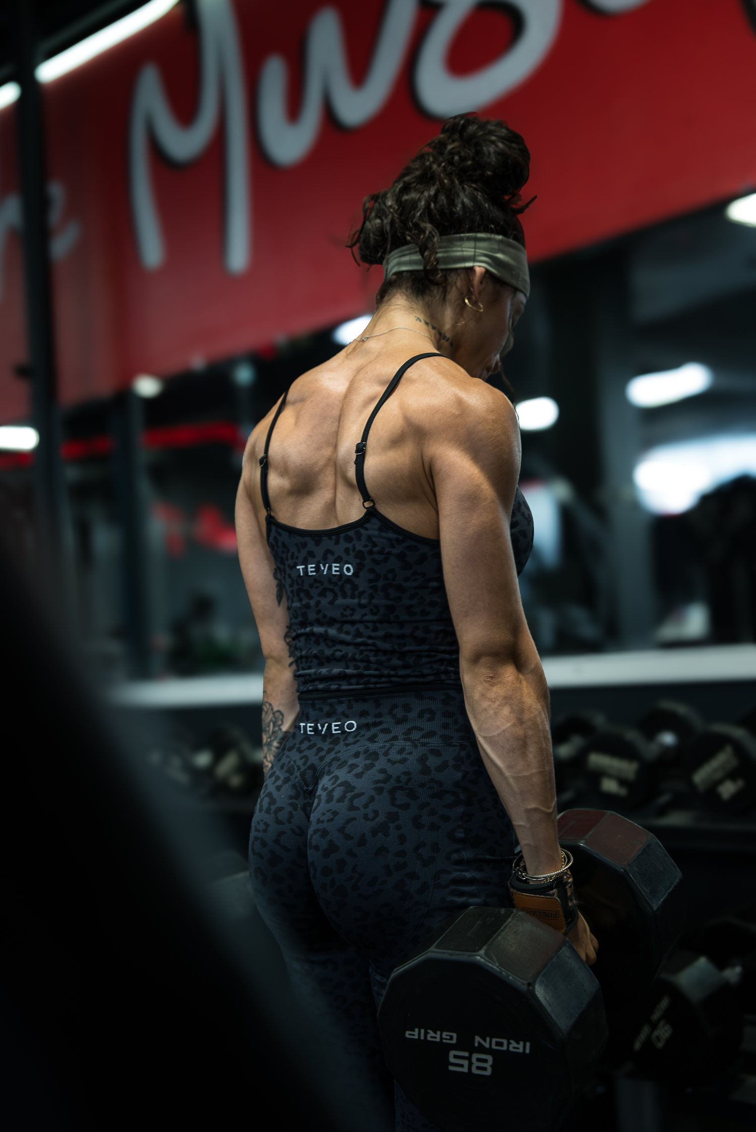 Roxy Cisneros from behind with a dumbbell in each hand, her back lit by the gym lights