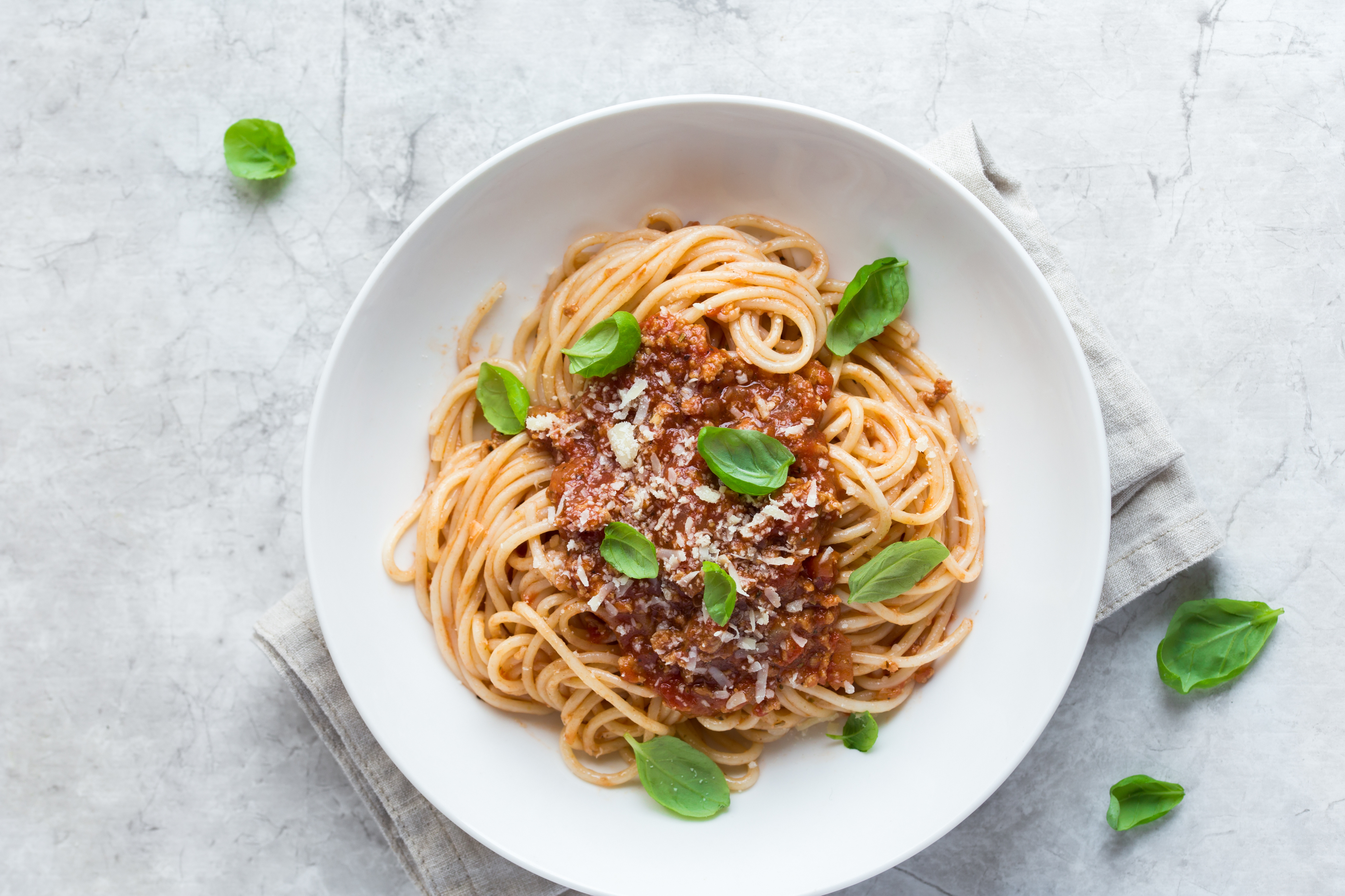 White bowl of spaghetti with tomato sauce, grated cheese, and fresh basil leaves on a cloth napkin.