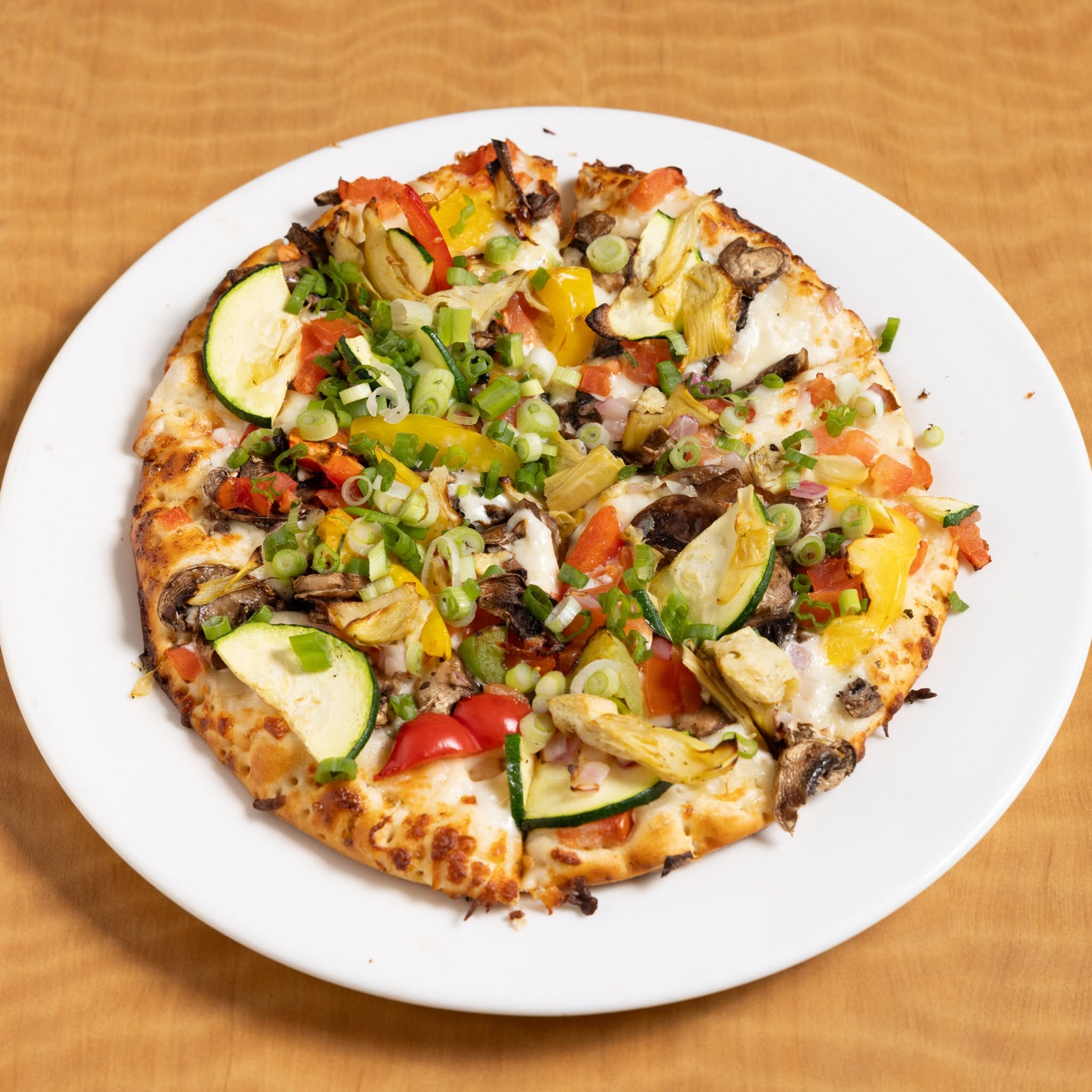 Vegetable pizza with mushrooms, zucchini, bell peppers, tomatoes, and green onions on a white plate.