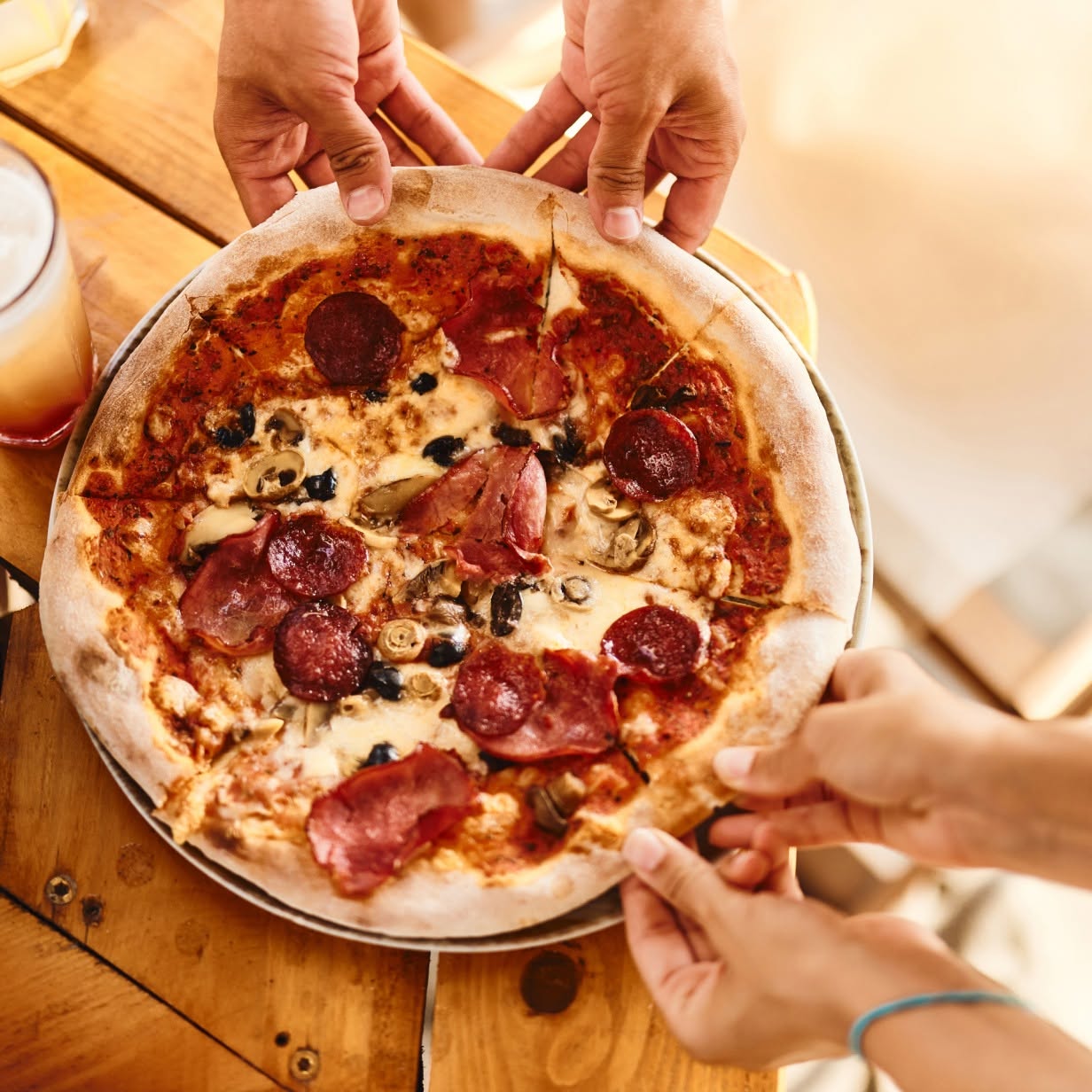 Hands holding a pepperoni and mushroom pizza on a wooden table with a drink nearby.