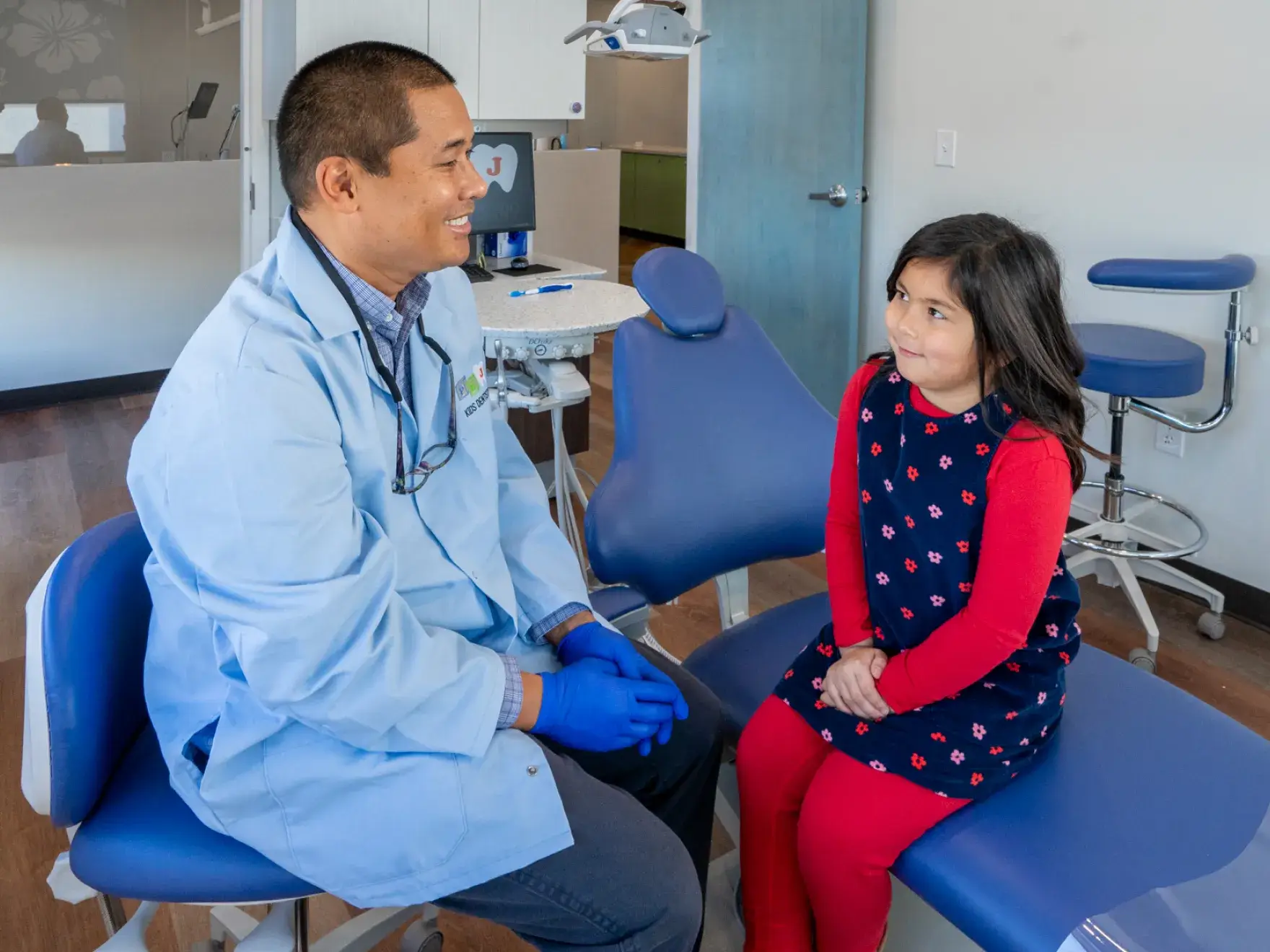 A little girl sitting on a blue chair next to a dentist.