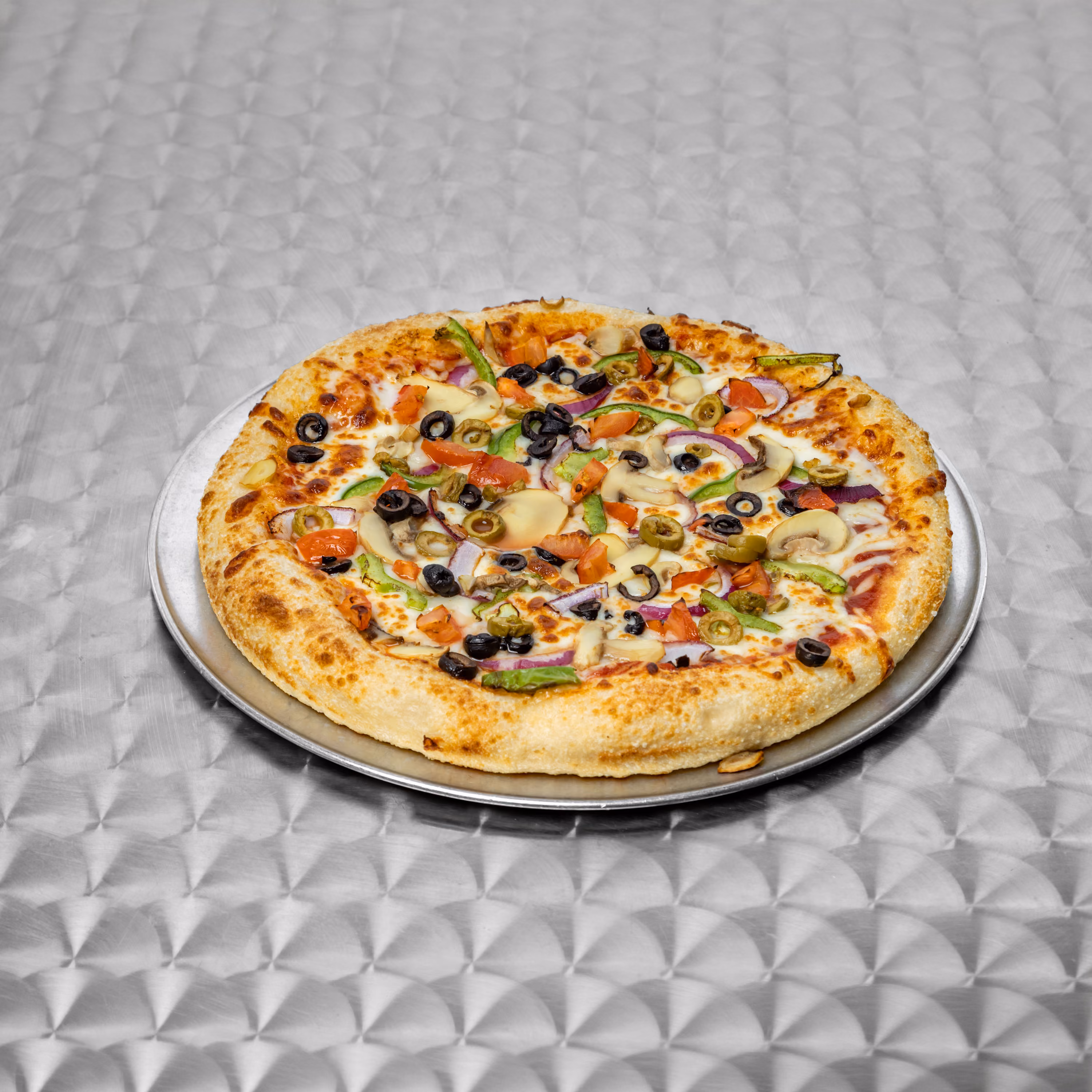 Vegetable pizza with olives, mushrooms, tomatoes, and peppers on a metal plate on a patterned table.