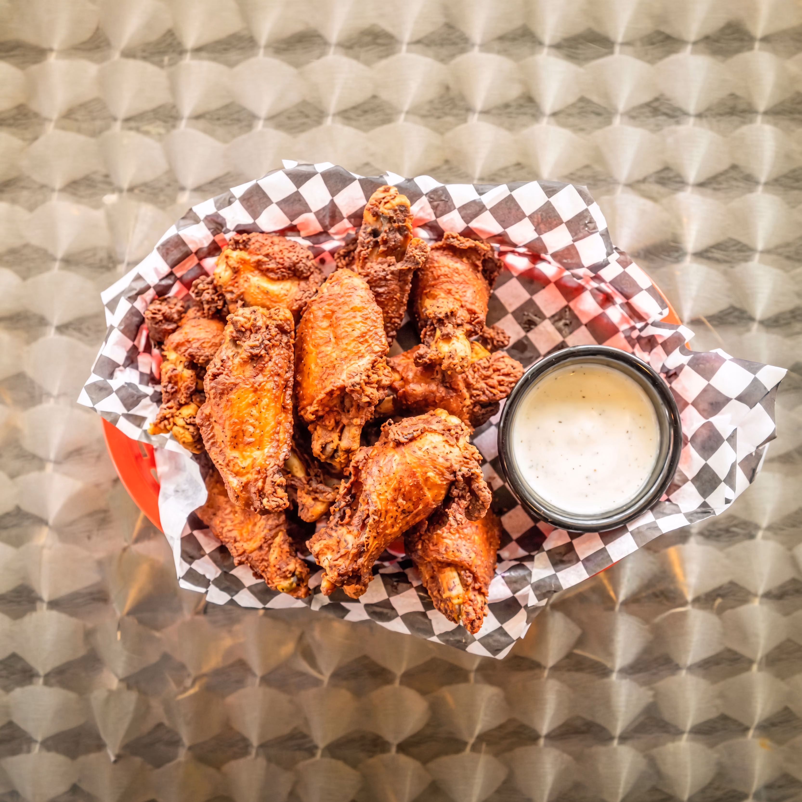 Basket of crispy fried chicken wings with a side of ranch dipping sauce on a metal table.