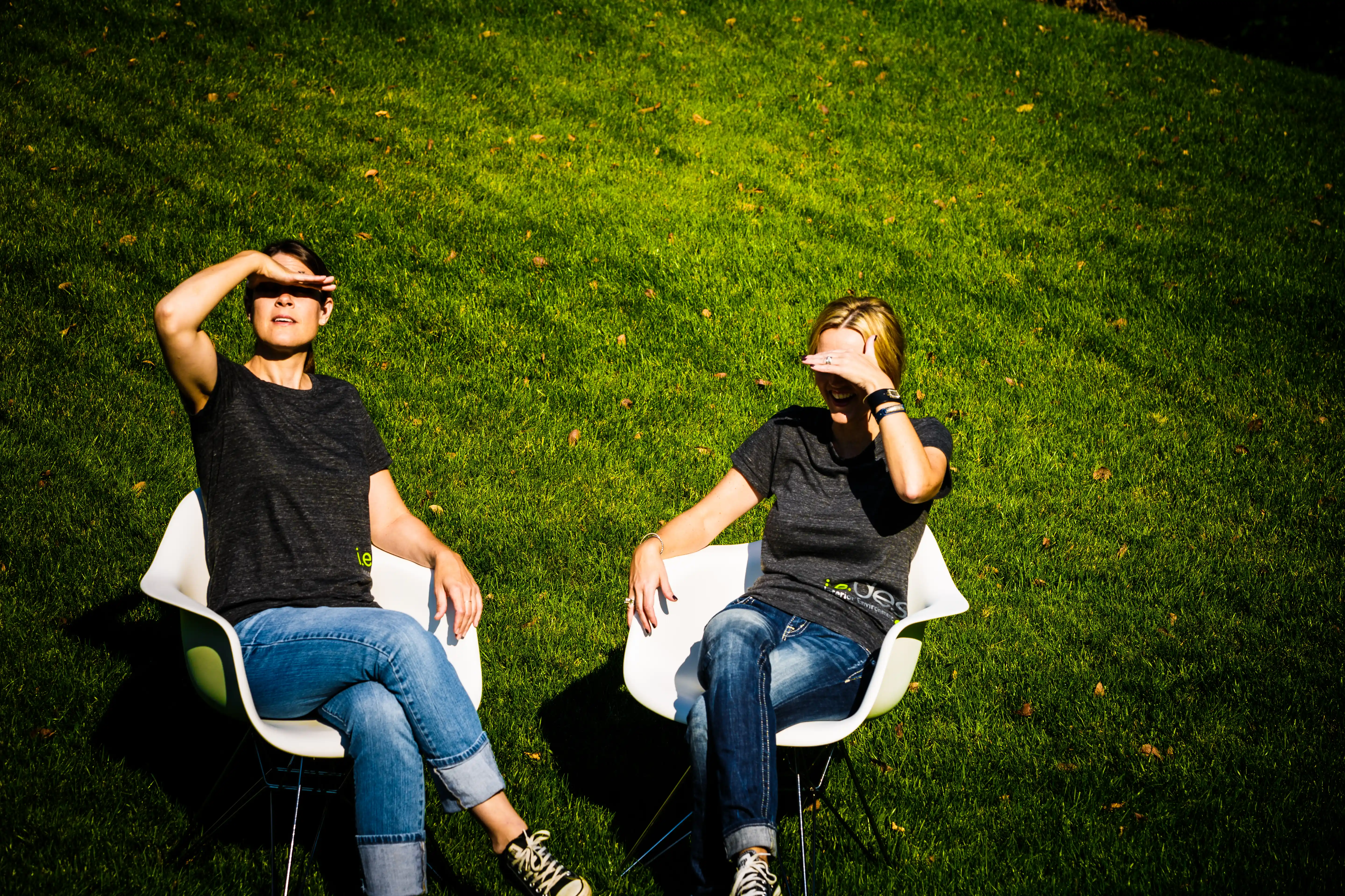 Two women, Angela and Jill, sit in white chairs on a sunlit lawn, shielding their eyes from bright light; a casual outdoor scene with a relaxed mood.
