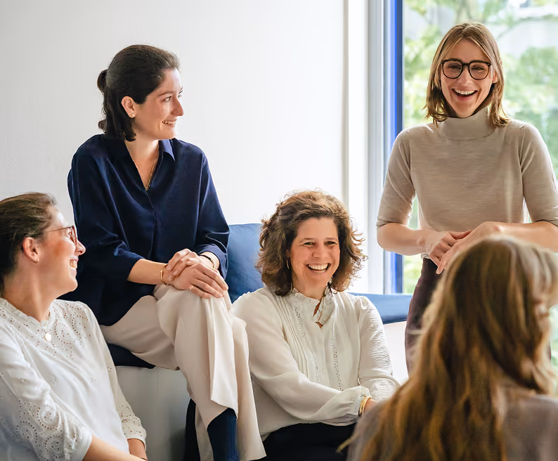 Frauen im Team tauschen sich in einem hellen Raum aus; eine Kollegin steht lächelnd neben der sitzenden Gruppe.