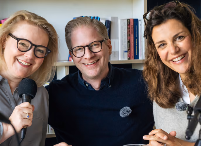 Drei Podcast-Gäste sitzen mit Mikrofonen vor einem Bücherregal.