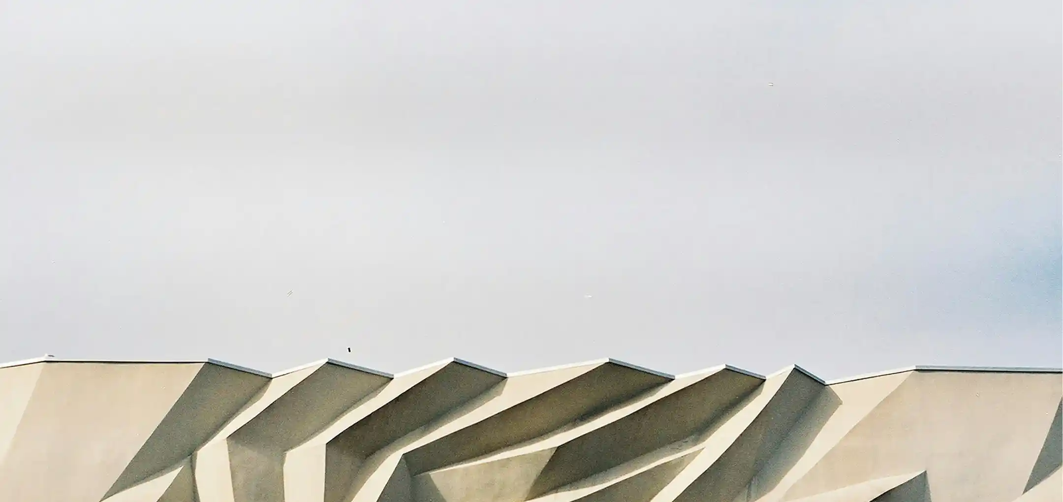Close-up of a building's architectural detail showing zigzagging concrete overhangs casting angular shadows against a light gray sky.