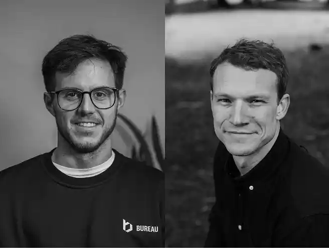 Black and white photo showing two men side by side. The man on the left wears glasses and a sweatshirt with a bureau logo, smiling with a blurred indoor background. The man on the right has short hair, wears a buttoned shirt, and smiles slightly outdoors with grass behind him.