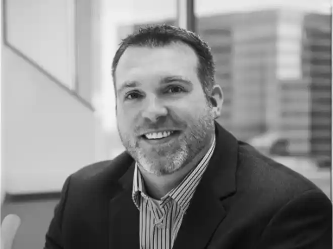 Black and white portrait of a smiling man with short hair and beard, wearing a striped shirt and dark blazer, sitting indoors with blurred office windows in the background.