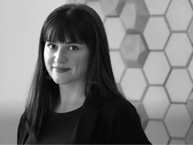 Black and white portrait of a woman with long dark hair and bangs, smiling slightly, wearing a dark top and jacket, standing in front of a hexagonal patterned wall.