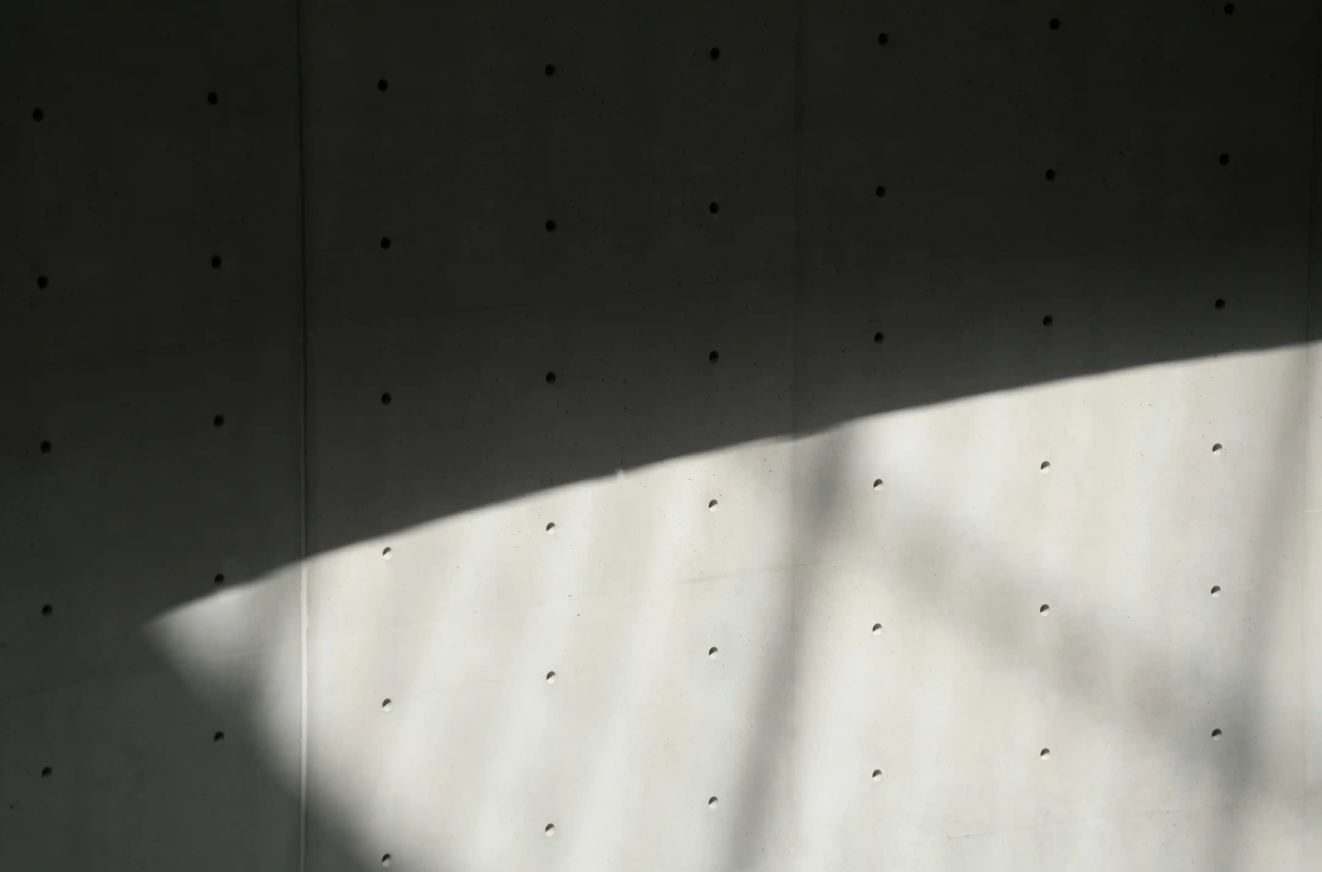 Concrete wall with a grid of evenly spaced small circular holes, partially illuminated by natural light casting soft shadows and shapes across the surface.