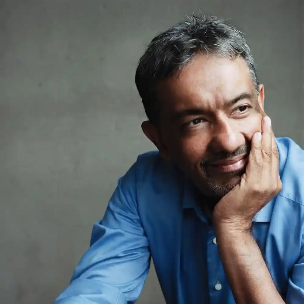 Man with short salt-and-pepper hair and a trimmed beard wearing a blue shirt resting his face on his hand, looking thoughtfully to the side against a plain gray background.