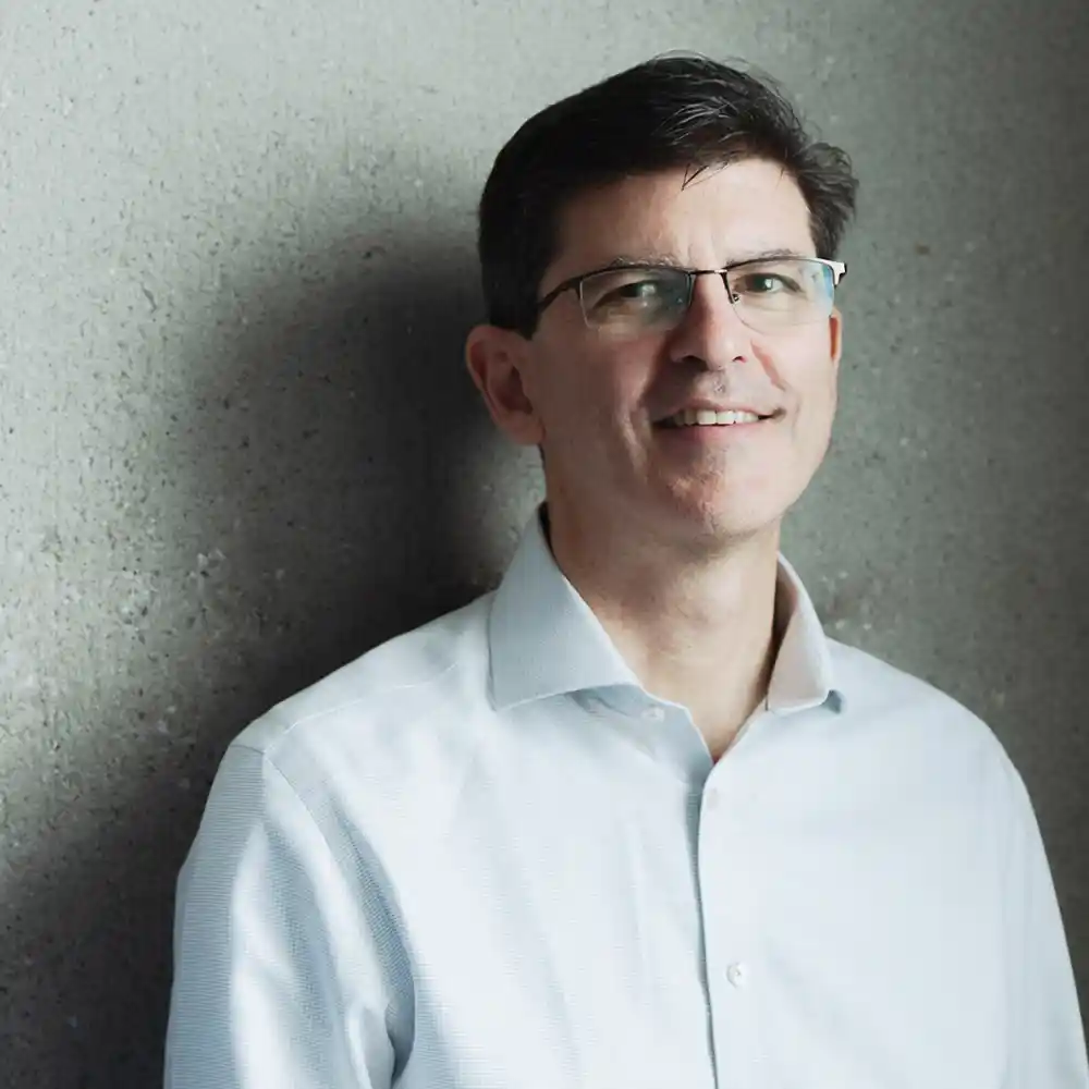 Smiling man with dark hair, glasses, and a light blue button-up shirt standing against a textured gray wall.
