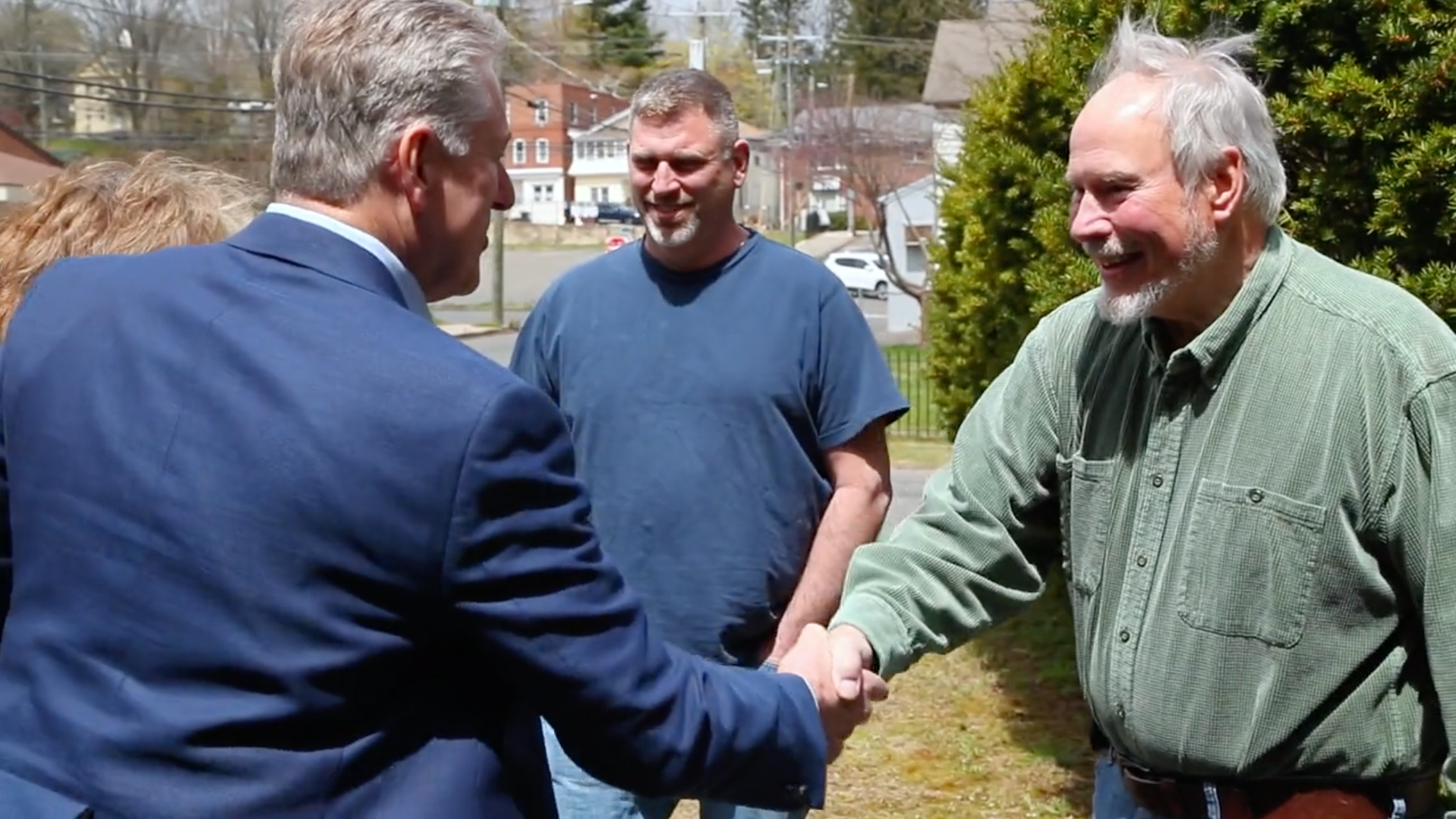 Two men shaking hands outside a manufacturing facility, with two colleagues looking on.