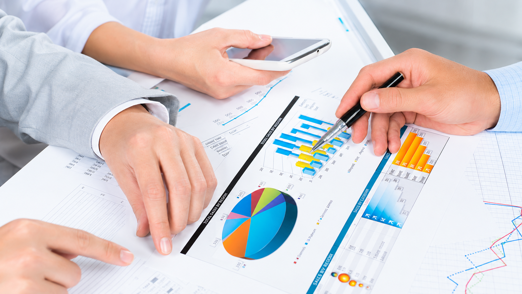 Two people reviewing printed sales charts and spreadsheets at a desk, one holding a phone.