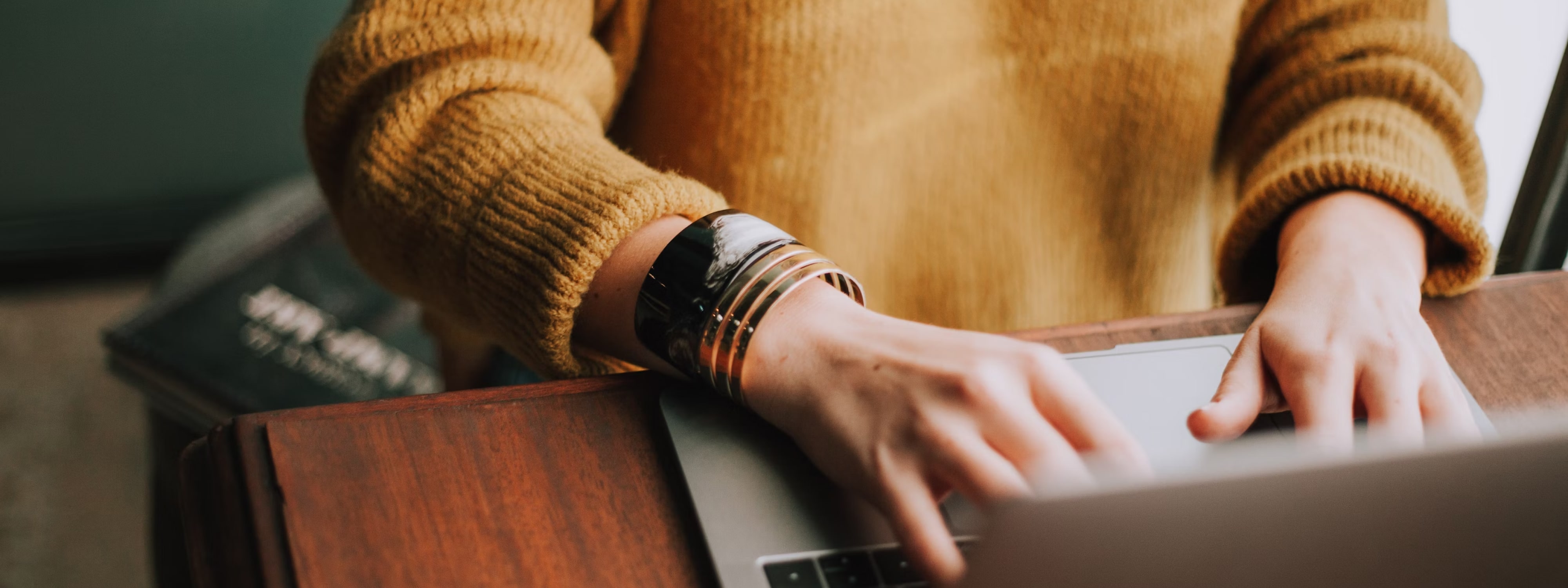 Close-up of a person's hands typing on a laptop, wearing a knit sweater and layered bracelets