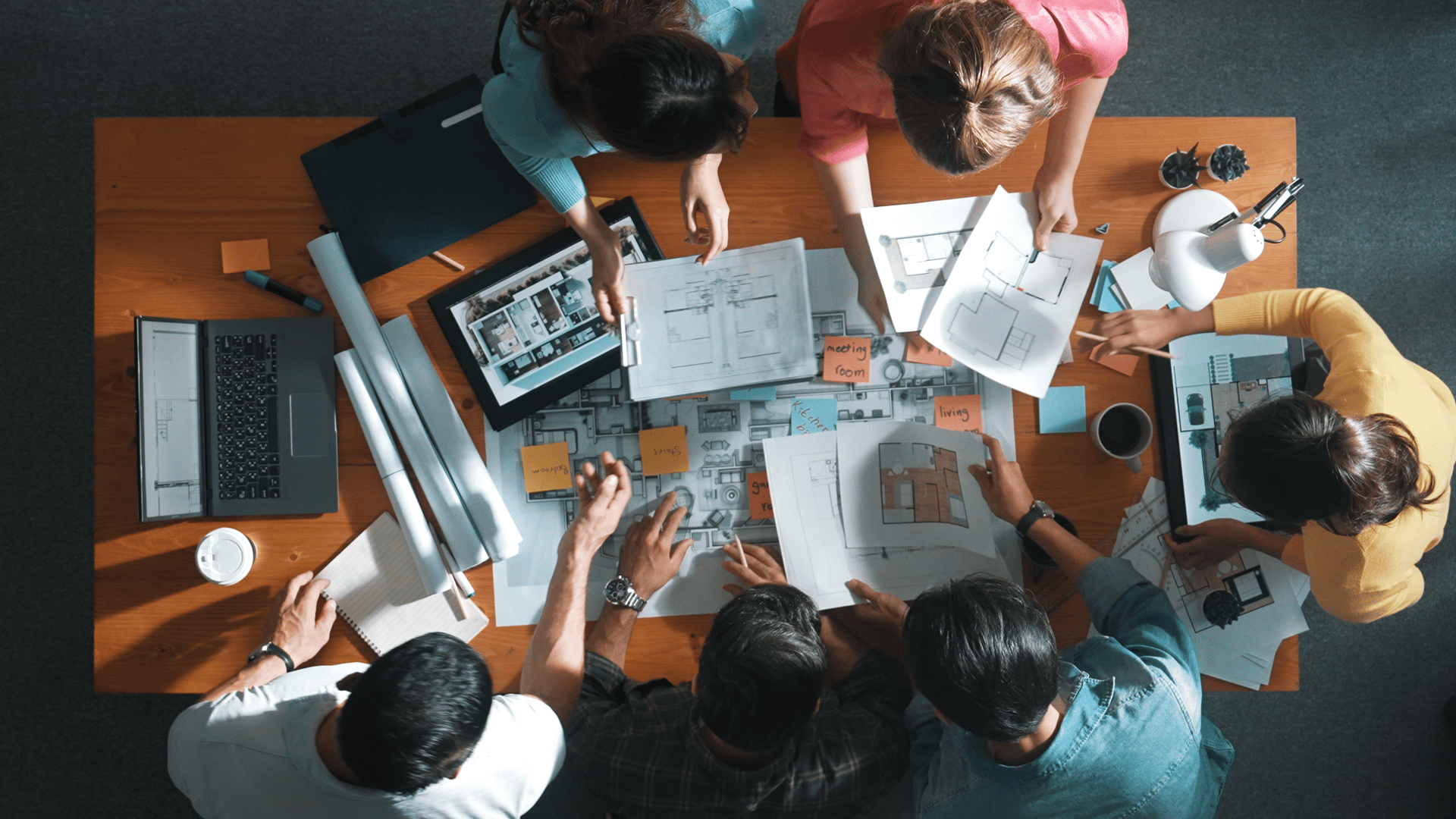 Overhead view of a diverse group collaborating around a table with blueprints, floor plans, and a laptop