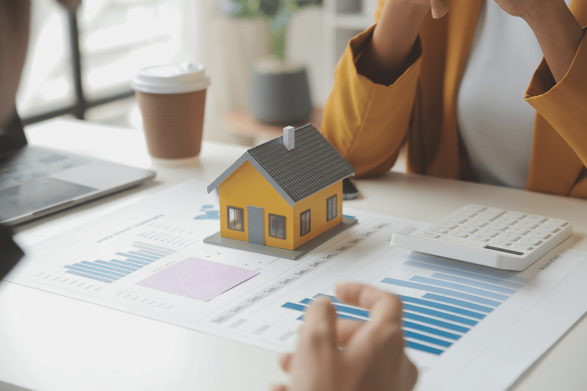 Person reviewing housing market data and a model house on a desk with charts and a calculator