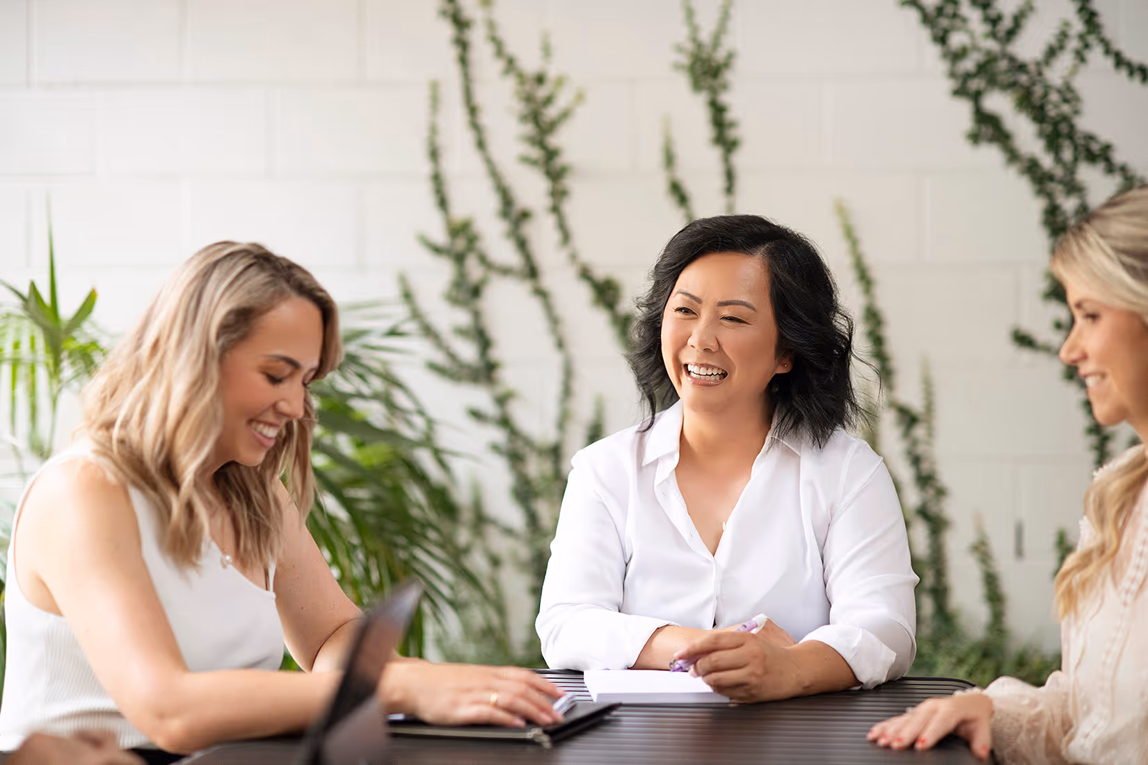 Three Catalyst Migration team members are sitting at a table, smiling and engaged in a friendly conversation with plants in the background.