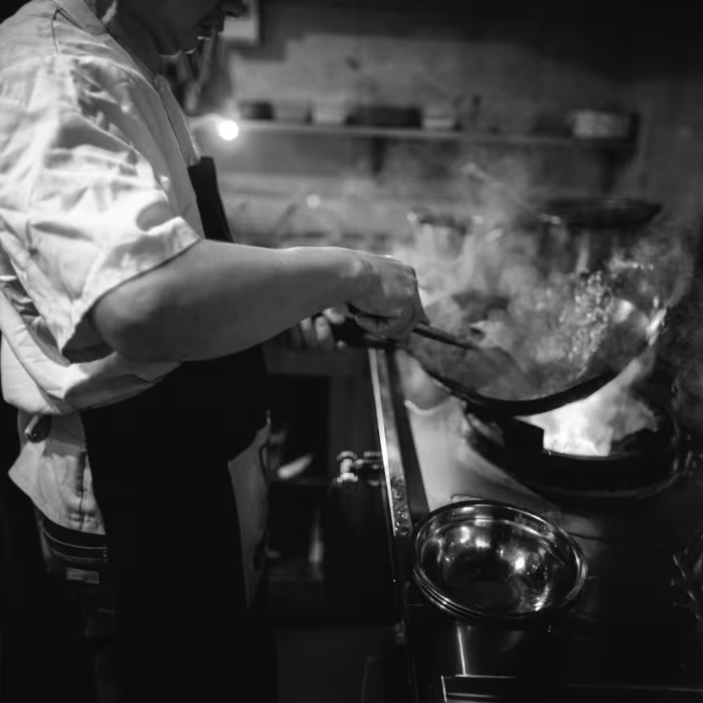 Chef in apron cooking with a wok over high heat on a stove in a kitchen emitting steam.