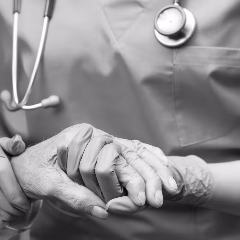 Healthcare professional wearing gloves gently holding an elderly person's hand with a stethoscope visible.
