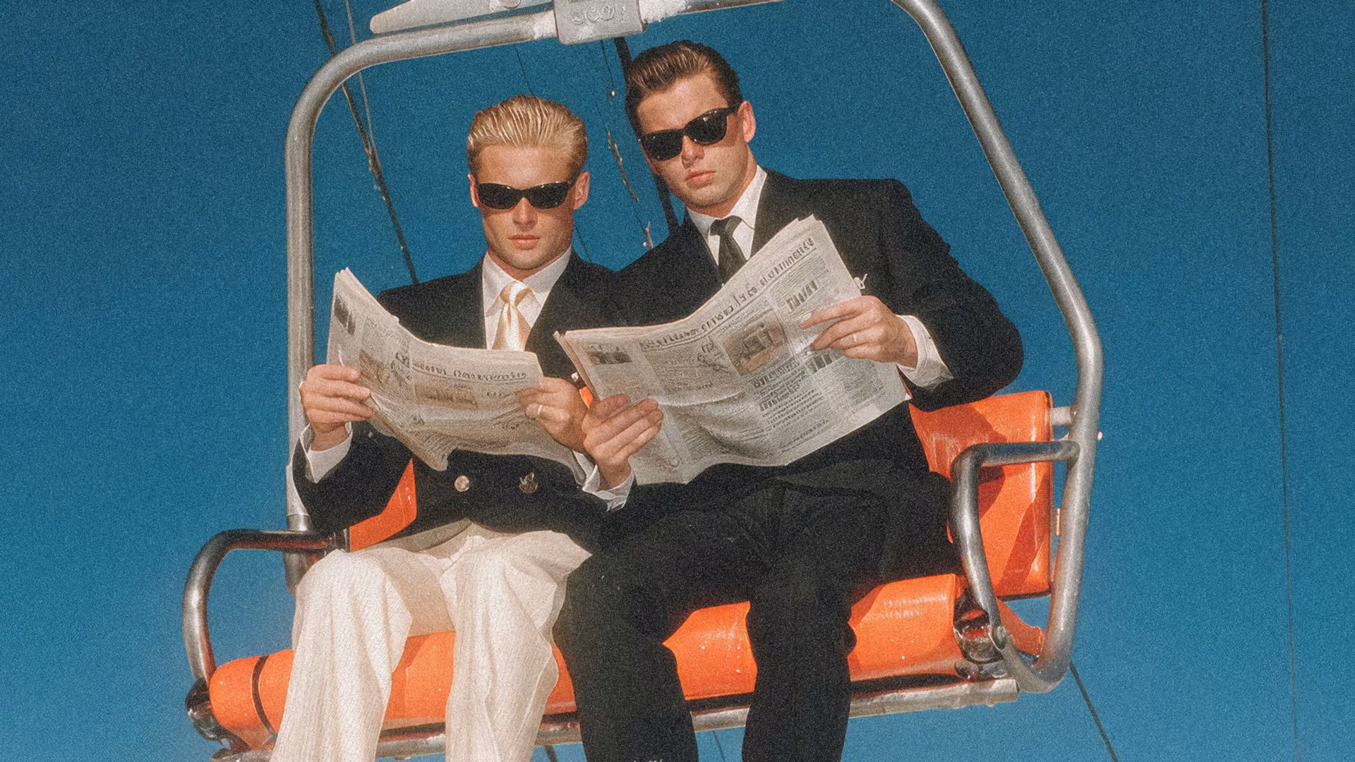 Two men in suits and sunglasses sitting on an orange chairlift reading newspapers against a blue sky.